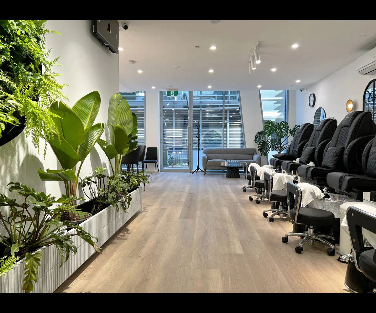 Room-length interior from alternate angle — plant wall and black pedicure chairs, grey sofa and windows at rear
