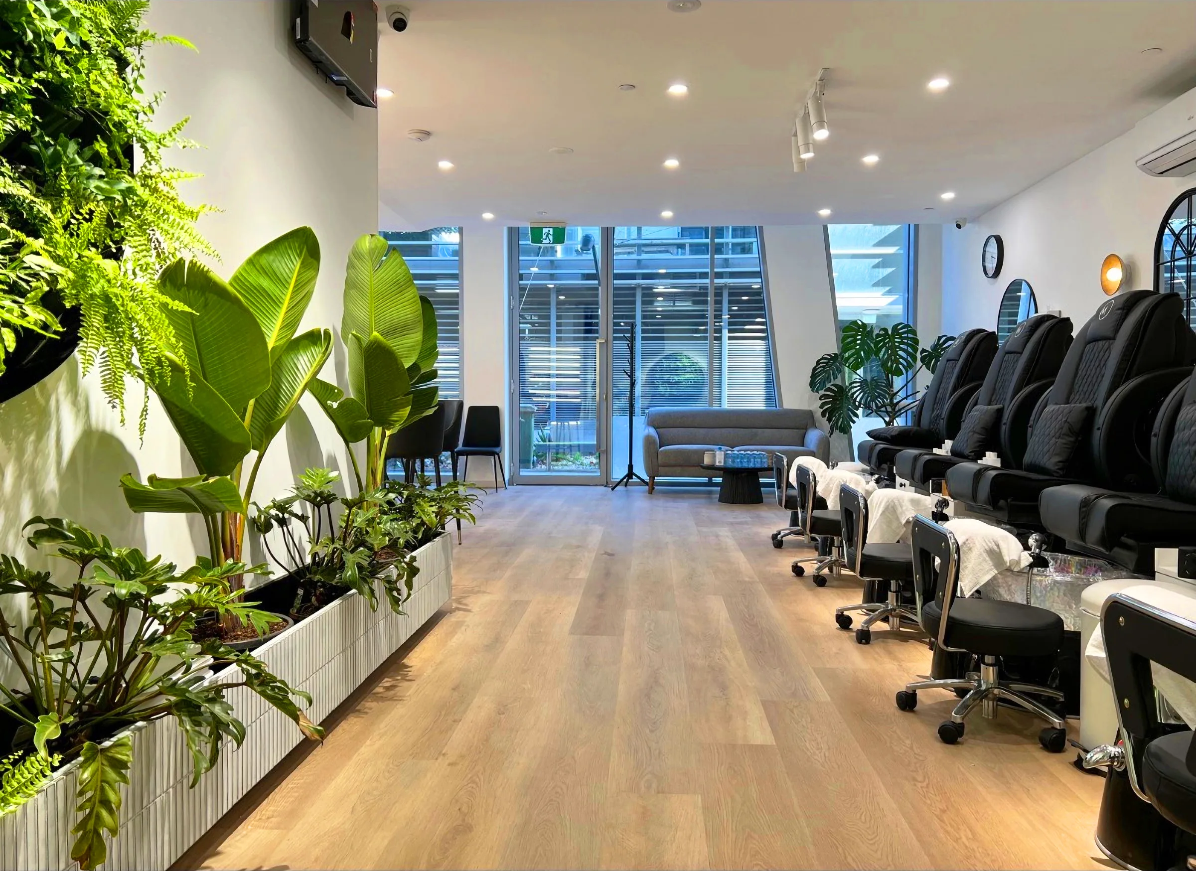 MONSTERA salon interior — room-length pedicure room with plant wall and black chairs receding to full-height windows