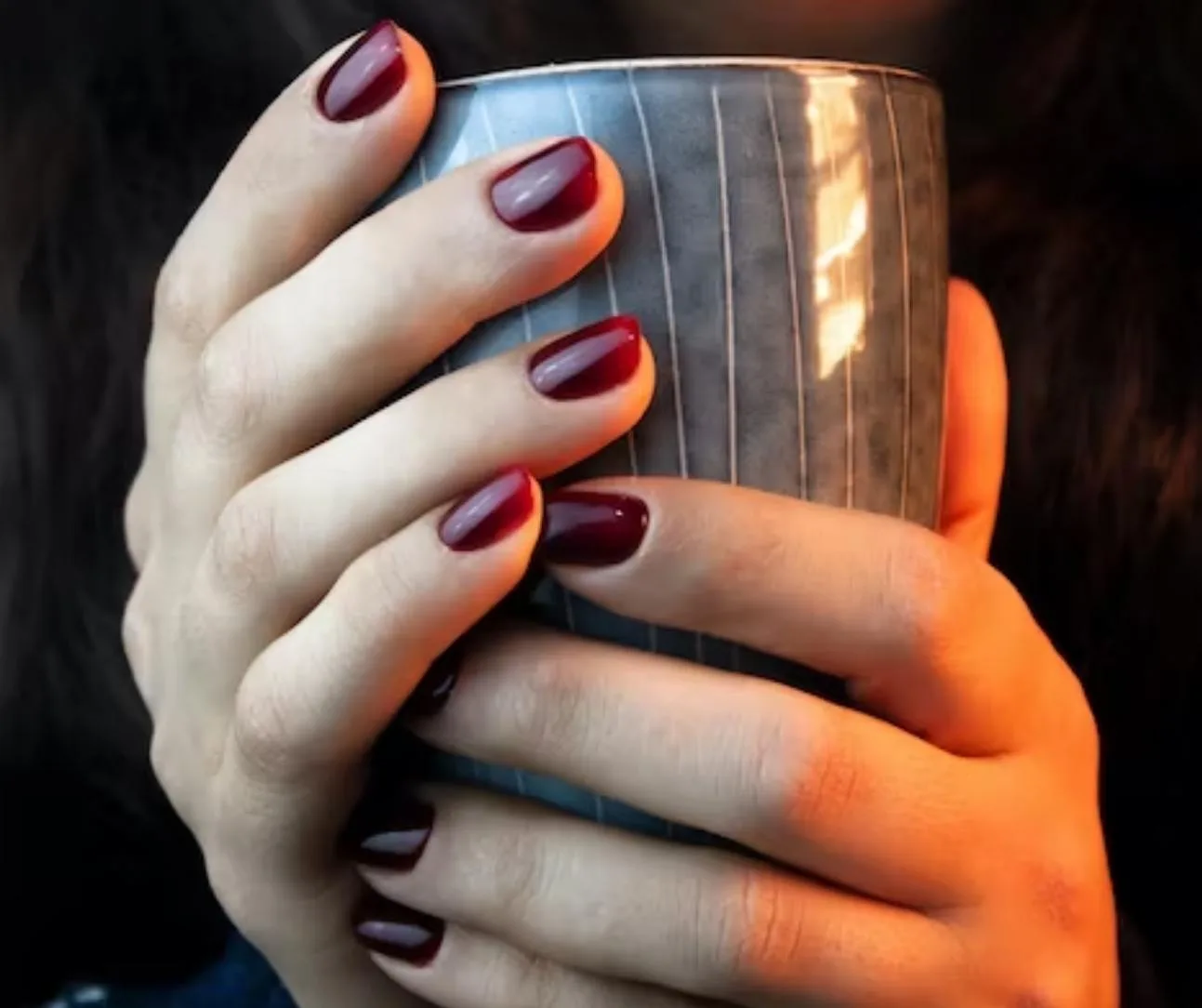 One hand cradling a grey cup with glossy deep-burgundy oval nails in warm light