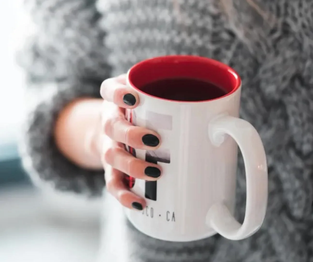 Cosy lifestyle image — hand with plain black nails holding a white and red mug against a grey knit