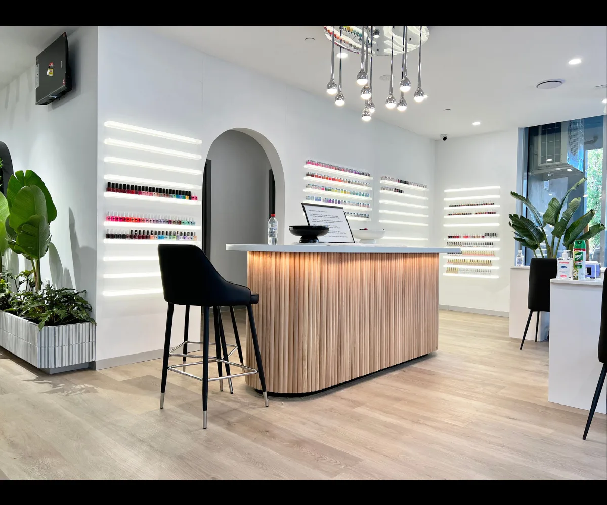 Reception close-up — curved wood-slat counter, LED nail-polish shelving, white arch, bar stool, plants